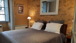 A bedroom with a large bed, a nightstand with a lamp, a framed picture on the wall, and a stone wall. - APARTMENT THE THREE FOUNTAINS (Sarlat-La-Canéda)