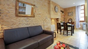 A living room with a stone wall, a brown leather sofa, a dining table with chairs, and a kitchen area with a clock and shelves. - APARTMENT THE THREE FOUNTAINS (Sarlat-La-Canéda)