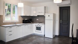 A modern kitchen with white cabinets, a gas stove, a microwave, and a refrigerator. There are two pendant lights, a window with a view of greenery, and a door to the right. - SUPERB RENT ANY COMFORT - POOL - BOULODROME - PRIVATE PARKING IN PEACE (Calenzana)