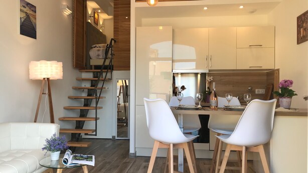 Un espacio habitable moderno con una escalera, una zona de comedor con sillas blancas, un sofá y una mesa con una revista y una planta en maceta.