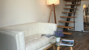 Una sala moderna con un sofá de cuero blanco, una mesa de centro de vidrio con una revista y flores, una escalera y una escalera que conduce a un dormitorio.