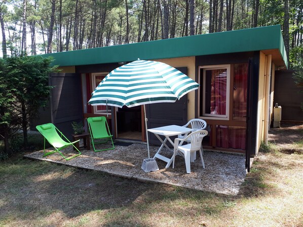 Une petite cabane composée de deux pièces, dotée d’un patio avec une table et deux chaises, un parasol et une chaise pliante verte.