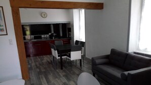 Ein modernes Wohnzimmer mit einem dunklen Sofa, einem weißen Tisch und einem Essbereich mit Stühlen. Im Hintergrund ist eine Küche mit roten Schränken und einer Wanduhr zu sehen.