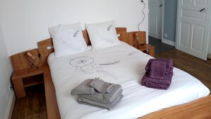 Ein ordentlich bezogenes Bett mit weißer Bettdecke, zwei Kissen, einer Nachttischlampe und einem Stapel Handtücher.