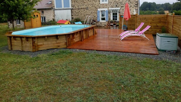 Ein Garten mit einem hölzernen Schwimmteich, einem Steinhaus und einer Holzterrasse mit Liegestühlen.