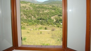 Vista a través de una ventana con marco de madera, sobre un paisaje montañoso con árboles dispersos y cielo despejado.