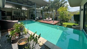 Une piscine extérieure moderne, dotée d’un patio couvert, entourée de plantes en pot et d’une pelouse bien entretenue.