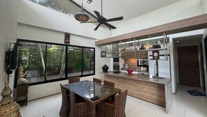 Une cuisine moderne dotée d’un îlot central, d’un coin repas avec des chaises en osier et d’une grande fenêtre offrant une vue sur la verdure.