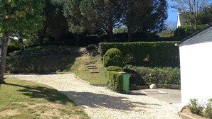 A gravel driveway leading to a white building with a wooden deck. There are trees and a hedge in the background.