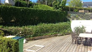 A wooden deck with a table and chairs, surrounded by well-maintained hedges and trees.