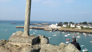 A stone cross on a rocky outcrop overlooking a harbor with boats and houses in the background.