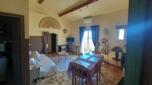 A room with a bed, a dining table with chairs, a small table with a lamp, a bookshelf, and a window with curtains. - Apartment Sea View Balcony & Shared Pool Air Conditioning (Hyères)