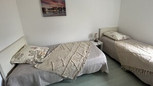 A small, well-lit bedroom with two single beds, a bedside table with a lamp, and a painting on the wall.