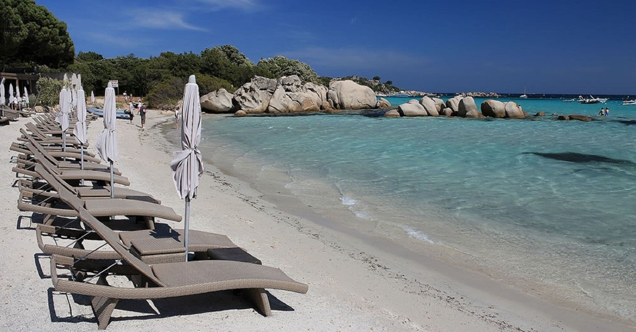 Ein Strand mit Reihen von Liegestühlen und Sonnenschirmen, klarem türkisblauem Wasser und Felsformationen im Hintergrund.