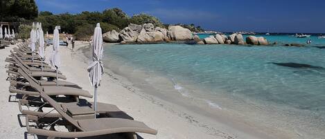 Ein Strand mit Reihen von Liegestühlen und Sonnenschirmen, klarem türkisblauem Wasser und Felsformationen im Hintergrund.