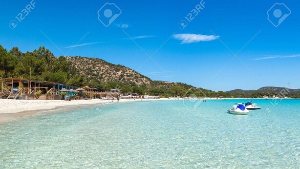 Une plage à l’eau turquoise et limpide, un bateau et une colline parsemée de bâtiments.