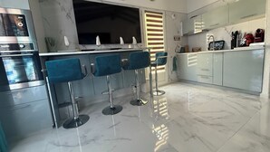Une cuisine moderne dotée d’un plan de travail en marbre, de quatre tabourets de bar bleus, d’un four encastré et d’une grande fenêtre avec des stores.
