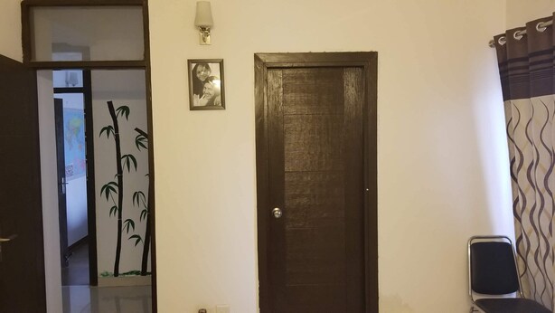 A room with a wooden door, a framed photo on the wall, a chair, and a decorative bamboo wall panel. - The most homely place in Noida (Sector 137)