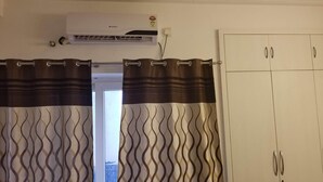 A room with a white wardrobe, a curtain with a wavy pattern, and an air conditioning unit on the wall. - The most homely place in Noida (Sector 137)