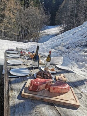 Une table de pique-nique en bois, installée à l’extérieur, avec des assiettes, des verres et des bouteilles de vin, entourée de neige et d’arbres.