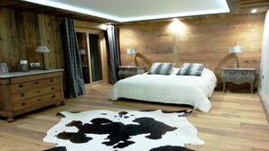 A bedroom with a wooden floor, a large bed with white bedding, two bedside tables with lamps, a wooden dresser, and a cowhide rug. - Exceptional "Frachette" Chalet (Praz-Sur-Arly)
