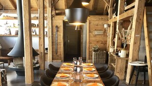 A long wooden dining table set with plates, glasses, and napkins, surrounded by grey upholstered chairs in a rustic wooden interior with exposed beams and a large chimney. - Exceptional "Frachette" Chalet (Praz-Sur-Arly)
