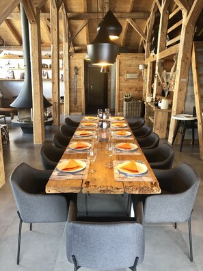 Una larga mesa de comedor de madera dispuesta con platos, vasos y servilletas, rodeada de sillas tapizadas grises en un interior rústico de madera con vigas a la vista y una gran chimenea.
