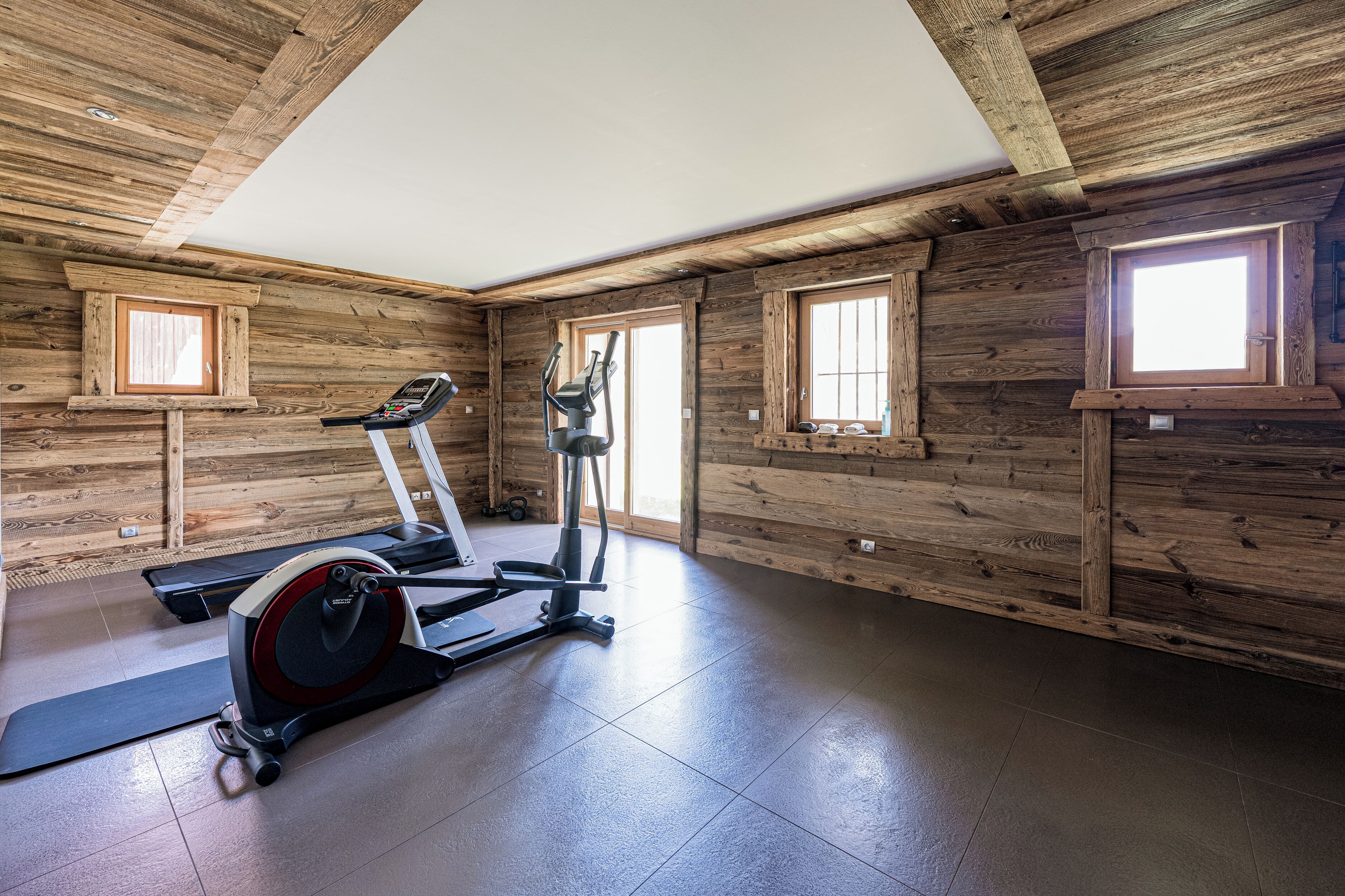 A gym with a treadmill, elliptical, and weights, wooden walls, and large windows.