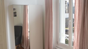 A room with wooden flooring, a window with pink curtains, a chair, and a mirror reflecting the room.