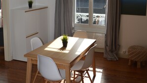 A dining area with a wooden table, two white chairs, a hanging lamp, a potted plant, and a view through a window with curtains.