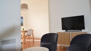 A modern living room with a flat-screen TV on a white cabinet, two dark armchairs, a dining area with a wooden table and chairs, and a pendant light.