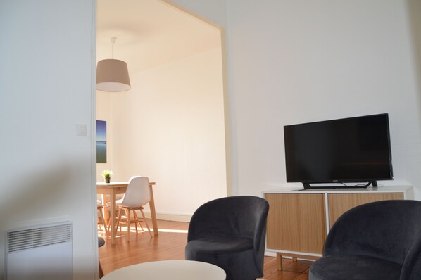 Una sala moderna con un televisor de pantalla plana sobre un mueble blanco, dos sillones oscuros, una zona de comedor con una mesa y sillas de madera, y una lámpara colgante.