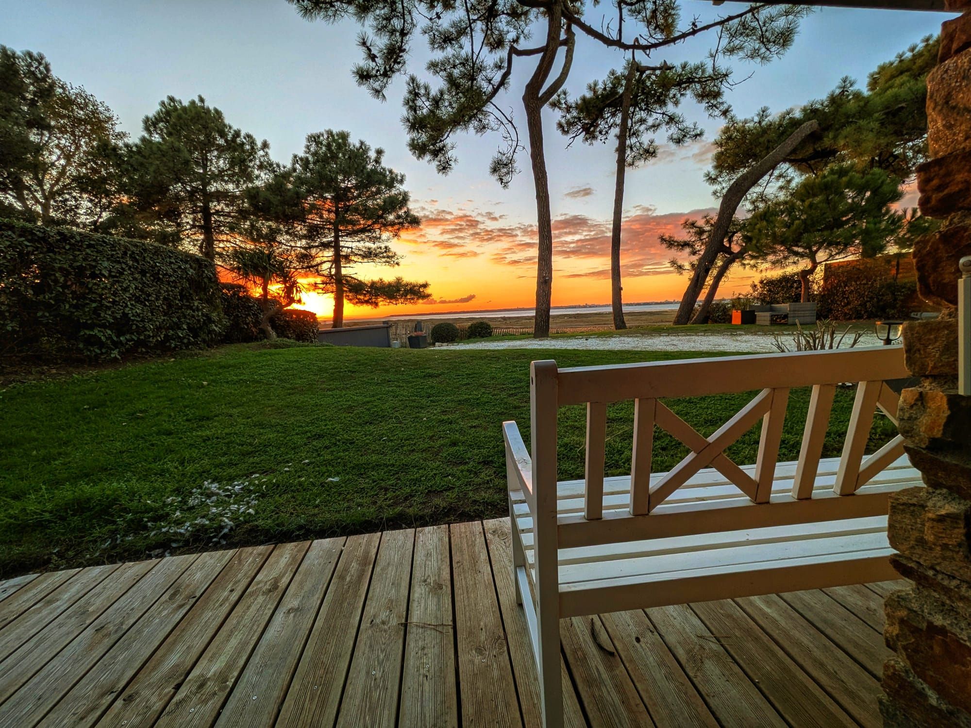 Een houten terras met een bankje, met uitzicht op een grasveld met bomen en een zonsondergang.