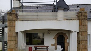 Uma área de estar coberta ao ar livre com um banco e cadeiras, um arco de pedra e uma janela com vista para um jardim.
