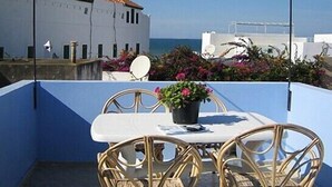 En balkon med et bord og stole, en potteplante og udsigt til havet og bygninger.