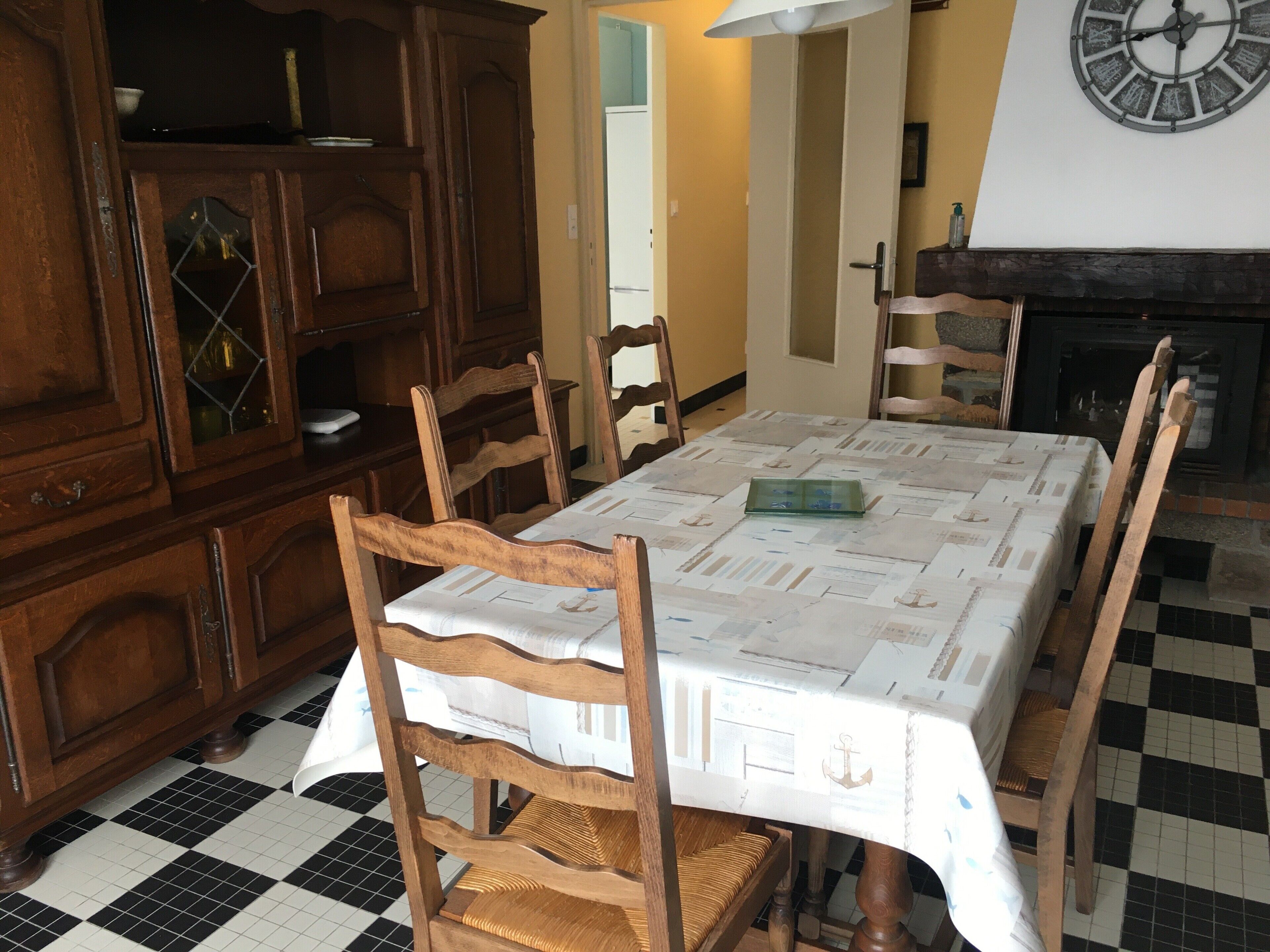 A dining area with a checkered floor, wooden chairs, a table covered with a patterned tablecloth, a wooden cabinet with glass doors, and a wall clock.