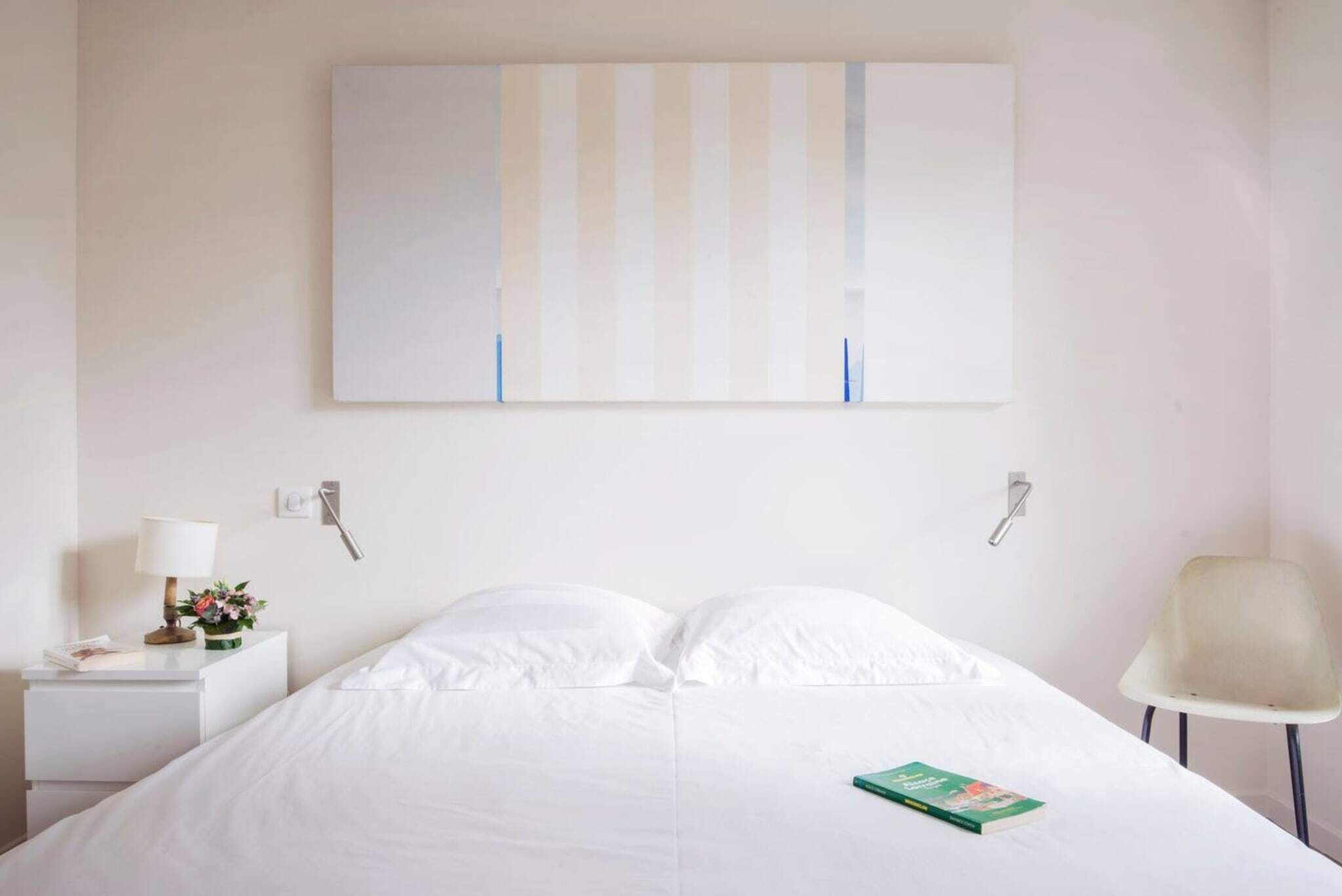 Een hotelkamer met een bed, nachtkastje met lamp, een stoel en een aan de muur hangend kunstwerk.