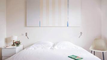 Een hotelkamer met een bed, nachtkastje met lamp, een stoel en een aan de muur hangend kunstwerk.