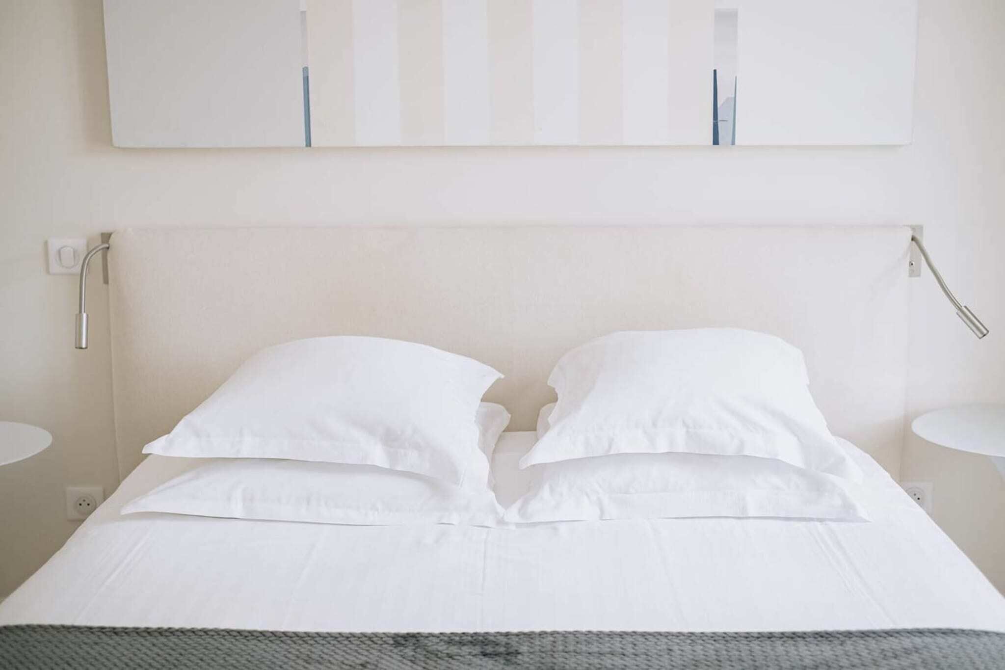 Un lit bien fait, avec du linge de lit blanc et une couverture grise, un mur blanc orné de rayures verticales, et une petite table de chevet blanche avec une lampe.