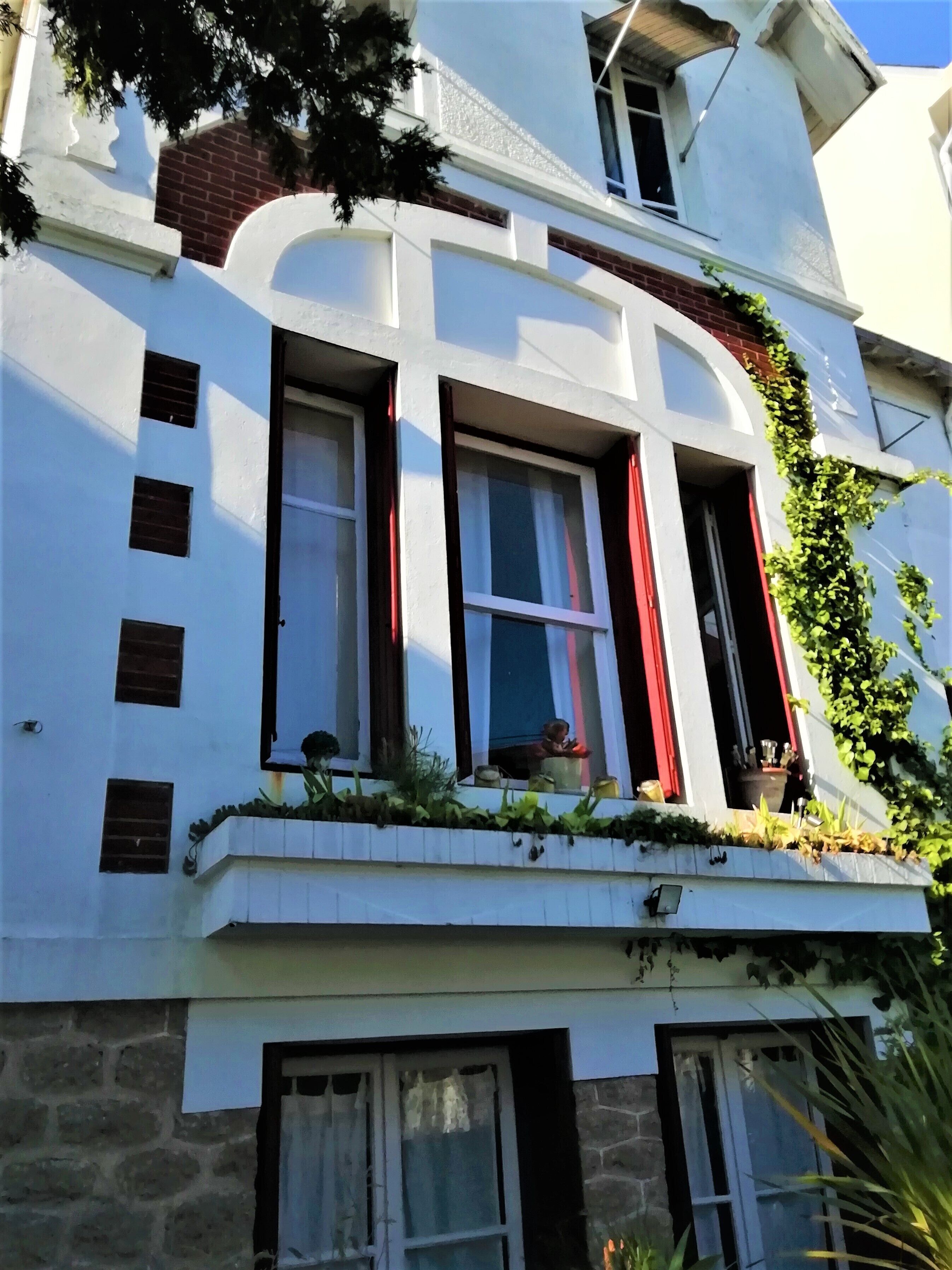 Um edifício de vários andares com fachada branca, várias janelas com cortinas vermelhas e uma varanda com plantas em vasos.