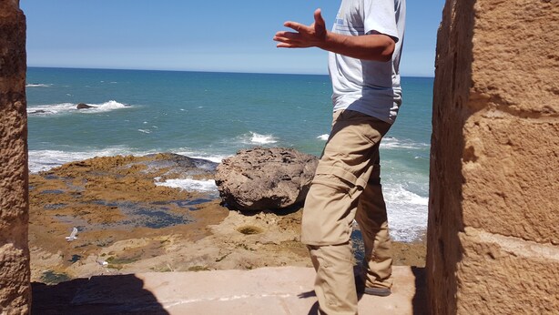 Un hombre de pie en una zona costera rocosa con olas rompiendo sobre la orilla.