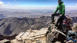 Una persona sentada sobre un saliente rocoso con un paisaje montañoso al fondo.