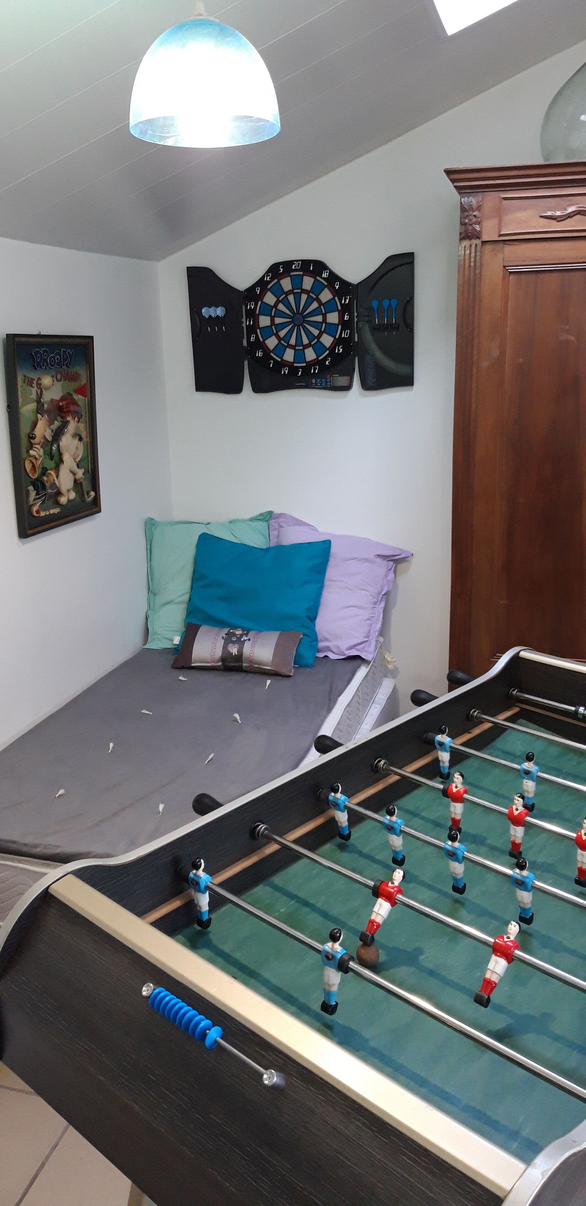 A room with a foosball table, a dartboard, a framed poster, and a bed with pillows.