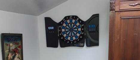 A room with a foosball table, a dartboard, a framed poster, and a bed with pillows.