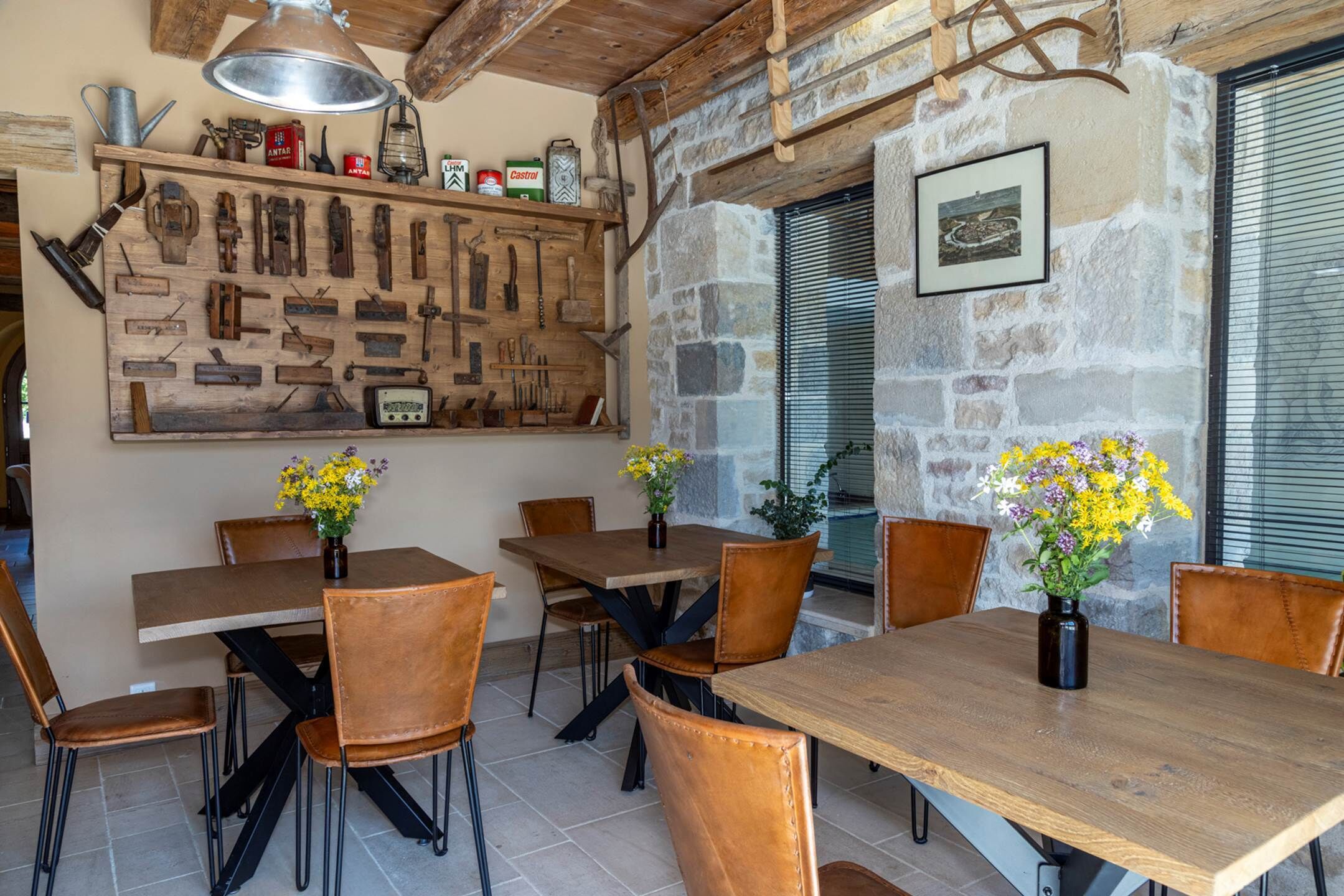A rustic dining area with wooden tables and chairs, a wall-mounted tool display, and a stone wall with a window.
