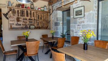 A rustic dining area with wooden tables and chairs, a wall-mounted tool display, and a stone wall with a window.
