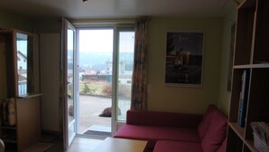 A living room with a sofa, a table, and a bookshelf. There is a door leading to a balcony with a view of buildings and a grassy area. - Apartment "Bellevue" Les Breuleux, Jura, Switzerland (Les Breuleux)