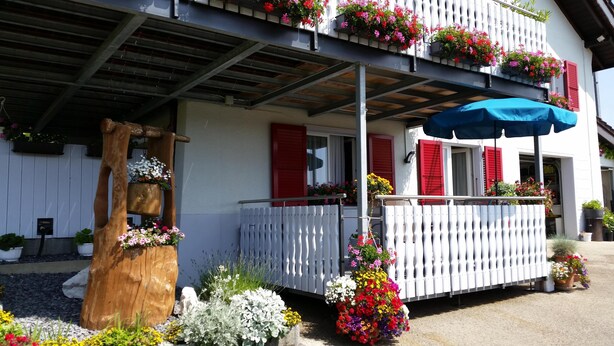 A two-story building with a balcony adorned with potted flowers and a blue umbrella. - Apartment "Bellevue" Les Breuleux, Jura, Switzerland (Les Breuleux)