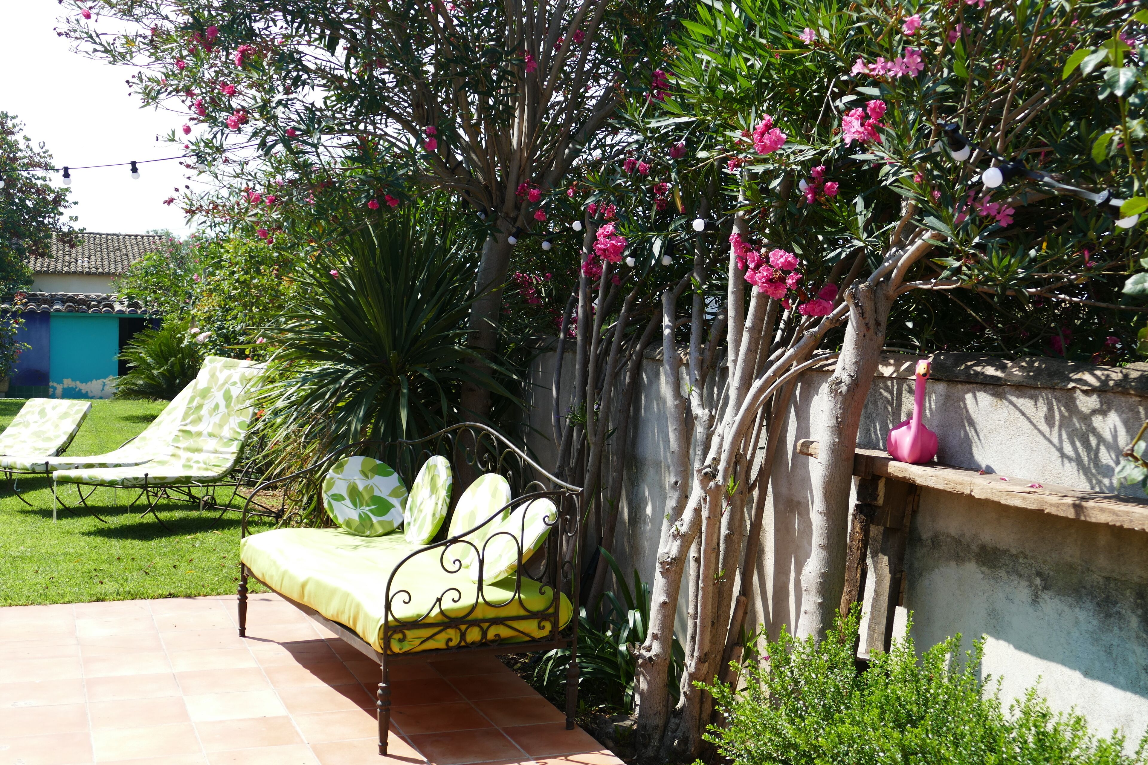 A patio with a metal bench and a small table, surrounded by lush greenery and flowering plants.