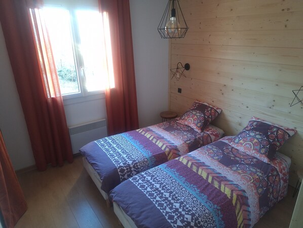 Et soveværelse med to senge, et vindue med gardiner, en hængende lampe og en træsenggavl.